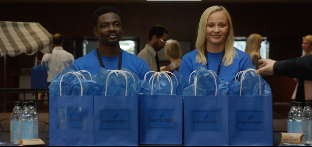 Still from AwareGO cybersecurity awareness video about conference risks. Two volunteers hand out blue gift bags that contain infected usb drives.